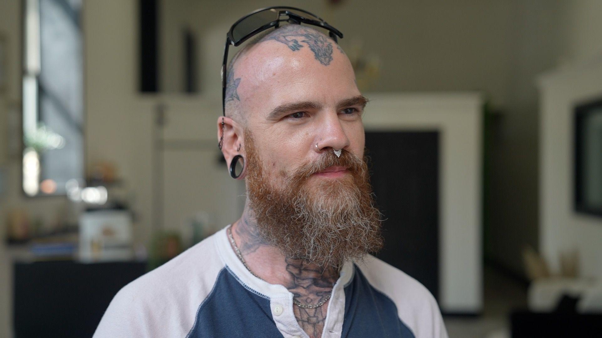 Adam Teague, calvo y barbudo, de 44 años, lleva gafas de sol en la cabeza, que está tatuada, junto con el cuello, y lleva dos piercings en la nariz