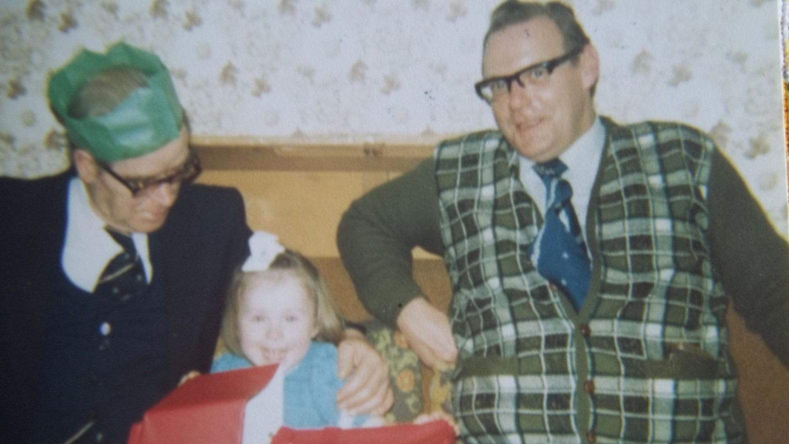 Fotografía familiar a color de la década de 1970. Anthony Littler, un hombre corpulento con gafas y una camiseta sin mangas a cuadros verdes con corbata azul, aparece sentado sonriendo en un sofá. A su izquierda, un hombre mayor con gafas y un gorro de fiesta de papel verde se inclina hacia una niña pequeña con un vestido azul que abre un gran regalo envuelto en papel rojo.