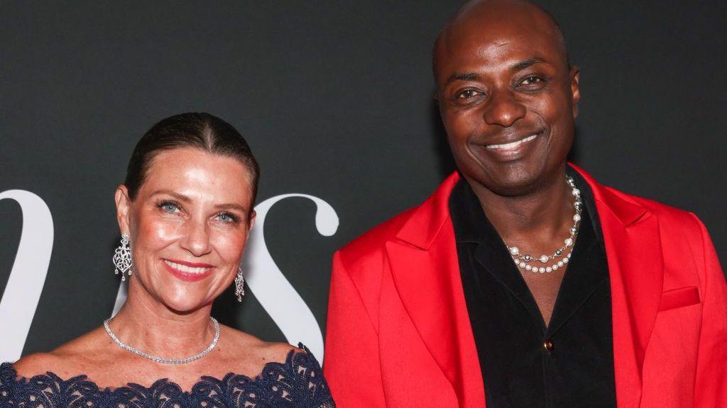 Marta Luisa (izquierda) y su esposo, el autodenominado chamán Durek Verrett (derecha), asisten a la primera edición de los Premios Gurus en el Hotel SLS de Beverly Hills, en Los Ángeles, el 10 de noviembre de 2024. Ambos sonríen a la cámara y lucen cadenas brillantes; ella lleva grandes aretes, mientras él viste camisa negra y chaqueta roja.