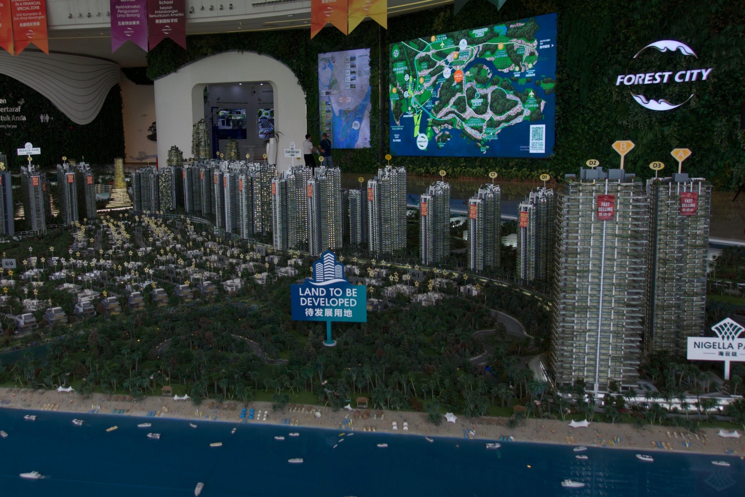 Un diorama de una ciudad, con un océano artificial de fondo, un gran cartel que dice "terreno para urbanizar" en un espacio verde artificial con árboles, y maquetas de rascacielos detrás, algunos con letreros que dicen "venta rápida". En la pared detrás del diorama hay una gran pantalla que muestra un mapa y un logotipo que dice "Forest City".