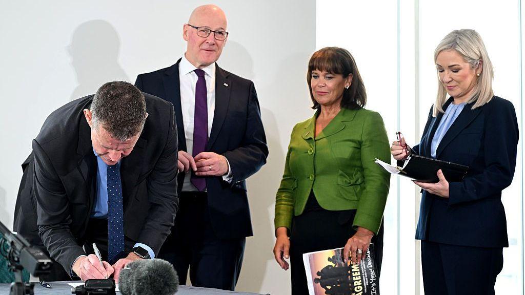 Cuatro personas, Michelle O'Neill, Mary Lou McDonald, Rhun ap Iorwerth y John Swinney, están de pie en una conferencia de prensa, mientras ap Iorwerth se inclina para firmar un documento.