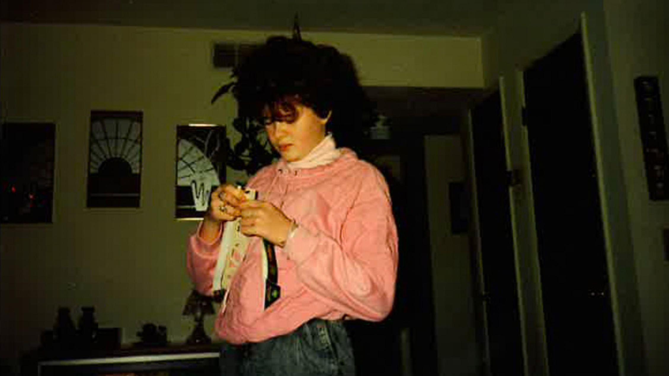 Christa Pike, fotografiada cuando era adolescente. Tiene el pelo castaño rizado y lleva una blusa rosa y vaqueros. Mira algo que tiene en las manos; la habitación está oscura.