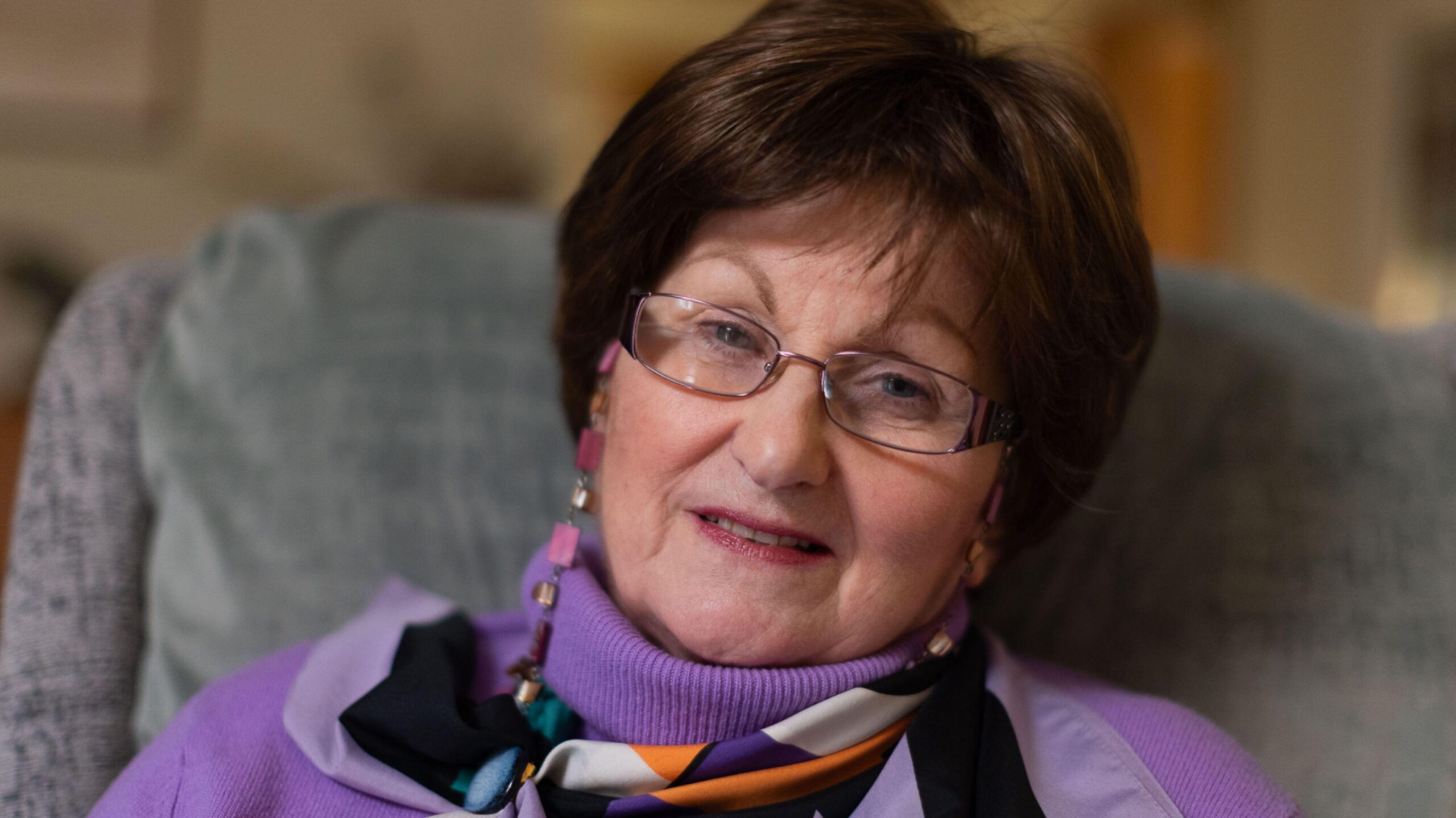 Una anciana de pelo corto y oscuro. Lleva gafas, un jersey morado y una bufanda colorida. Está sentada en un sillón gris, sonriendo a la cámara.