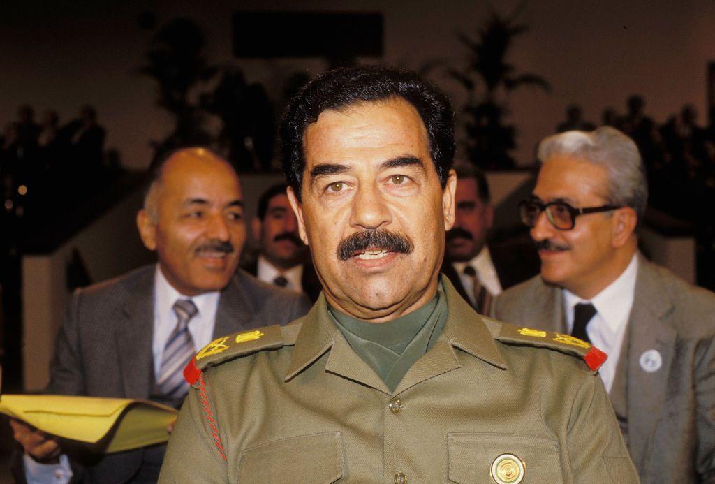 El expresidente iraquí Saddam Hussein asiste a un acto oficial vistiendo un uniforme militar verde oliva, con varios asistentes visibles al fondo.