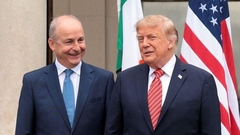 A la izquierda, Micheál Martin, con traje y corbata azules. A su derecha, Donald Trump, con traje oscuro y corbata roja. Detrás de ellos ondean las banderas irlandesa y estadounidense.