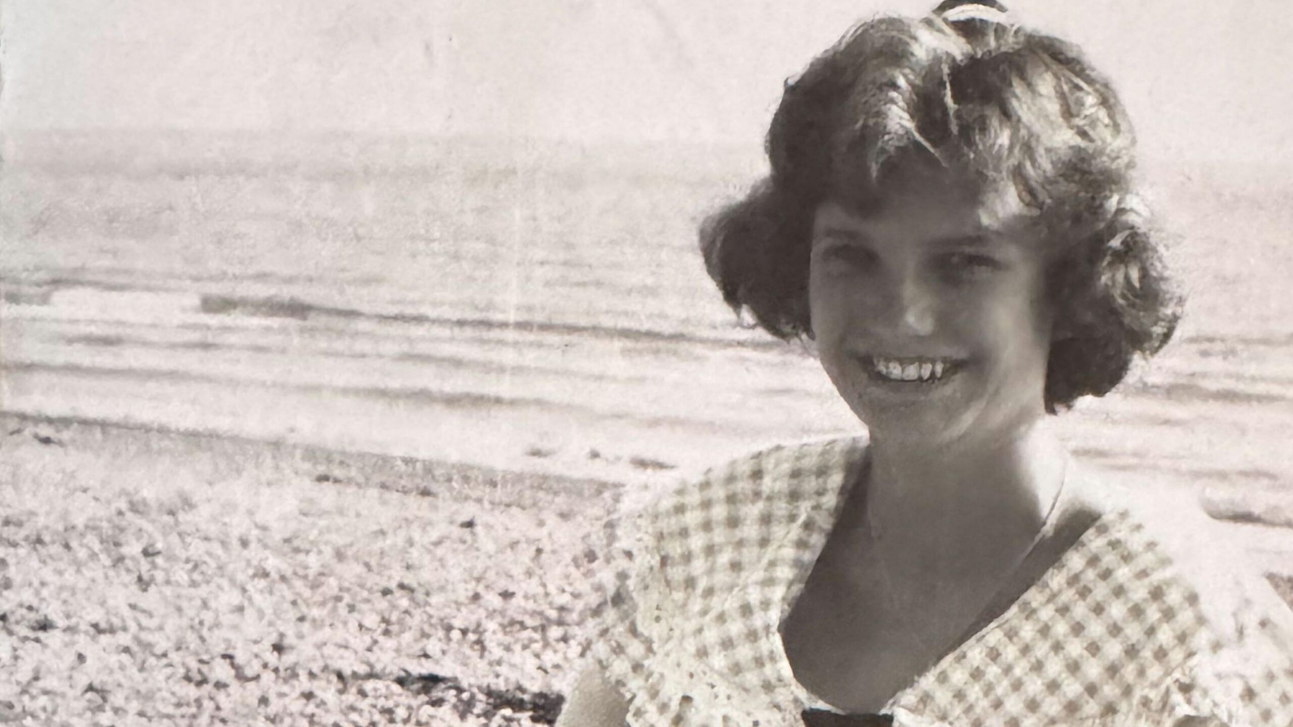 Fotografía en blanco y negro de una joven con un vestido de cuadros vichy. Está sentada en una playa de guijarros con el mar de fondo.
