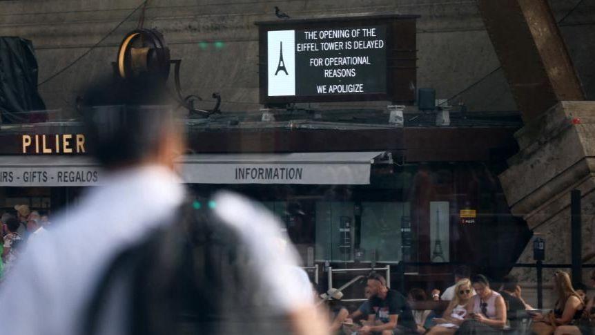 En primer plano de la fotografía, se ve desenfocado a un hombre con una mochila. La imagen muestra la entrada a la Torre Eiffel y un cartel que indica que está cerrada.