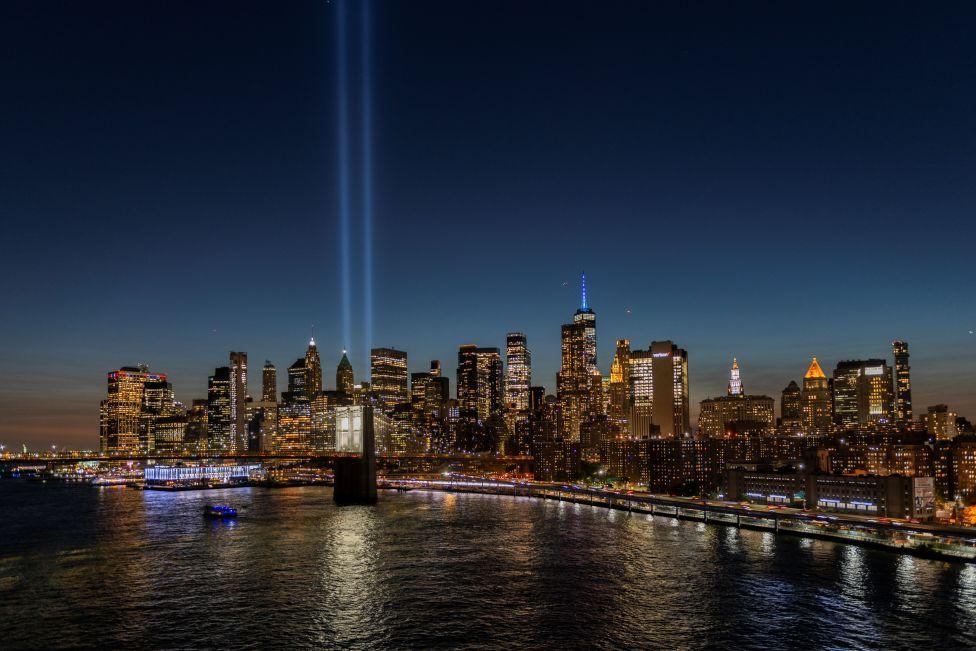 El horizonte de Manhattan por la noche, con los rascacielos iluminados contra un cielo oscuro. Dos haces de luz emanan del One World Trade Center para conmemorar las torres gemelas destruidas en 2001.