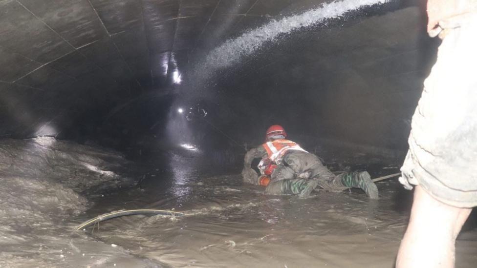 Un rescatista con uniforme verde y chaleco y casco anaranjados se arrastra por el suelo de una zona inundada