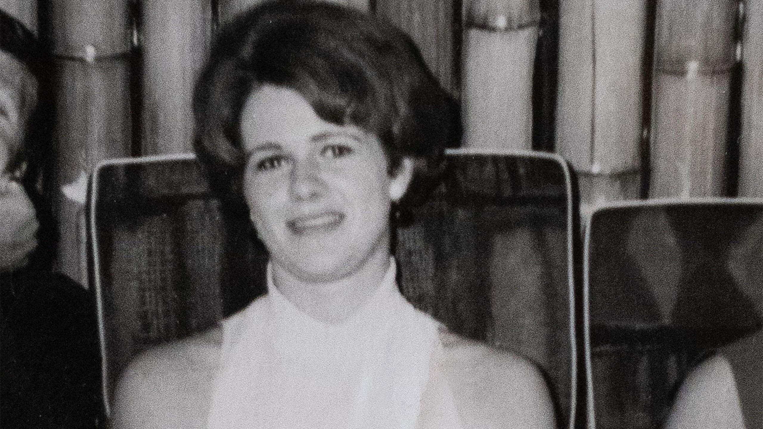 Fotografía en blanco y negro de una joven con una blusa blanca sin mangas. Está sentada en una silla frente a una cortina. 