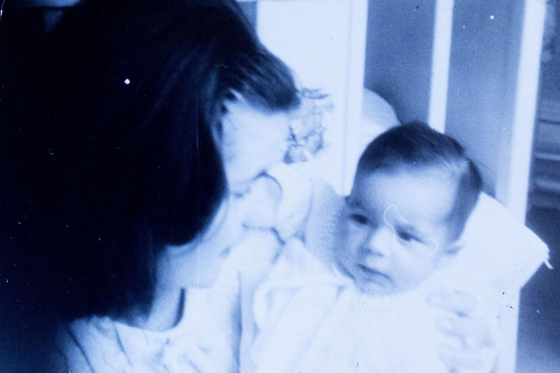 Una antigua fotografía en blanco y negro de una joven acunando a un bebé. Están agachados junto a una cuna. 