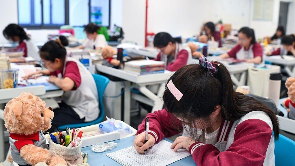 Una adolescente está sentada en un pupitre con la cabeza baja, mirando un papel. Escribe en él con un bolígrafo negro. Sobre su escritorio hay material escolar y un osito de peluche. Al fondo, se ven otros adolescentes sentados en sus pupitres, separados entre sí, en un aula.