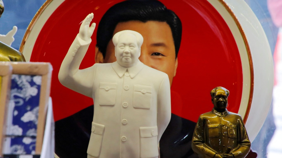 Dos figuras de Mao Zedong colocadas frente a un plato decorativo con una imagen sonriente de Xi Jinping. La figura más grande cubre gran parte del rostro de Xi.