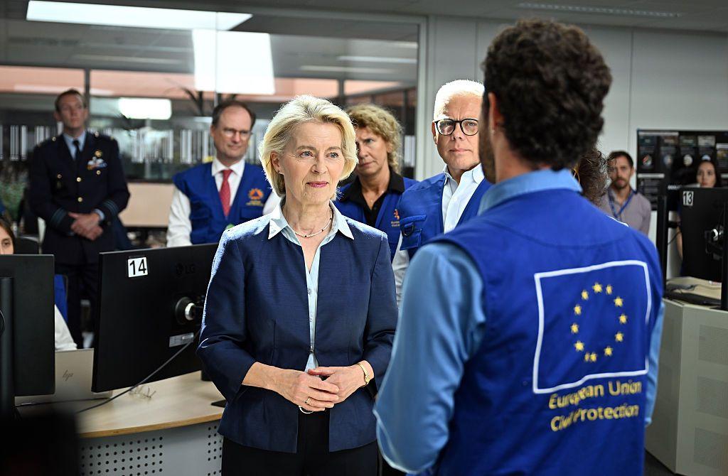 La presidenta de la Comisión Europea, Ursula von der Leyen (i), en actitud de escucha frente a una persona de espaldas a la cámara. Esa persona viste un chaleco azul con la bandera de la UE y el lema "European Union Civil Protection" en la espalda. Hay otras personas tras Von der Leyen en la oficina donde discurre la escena. 