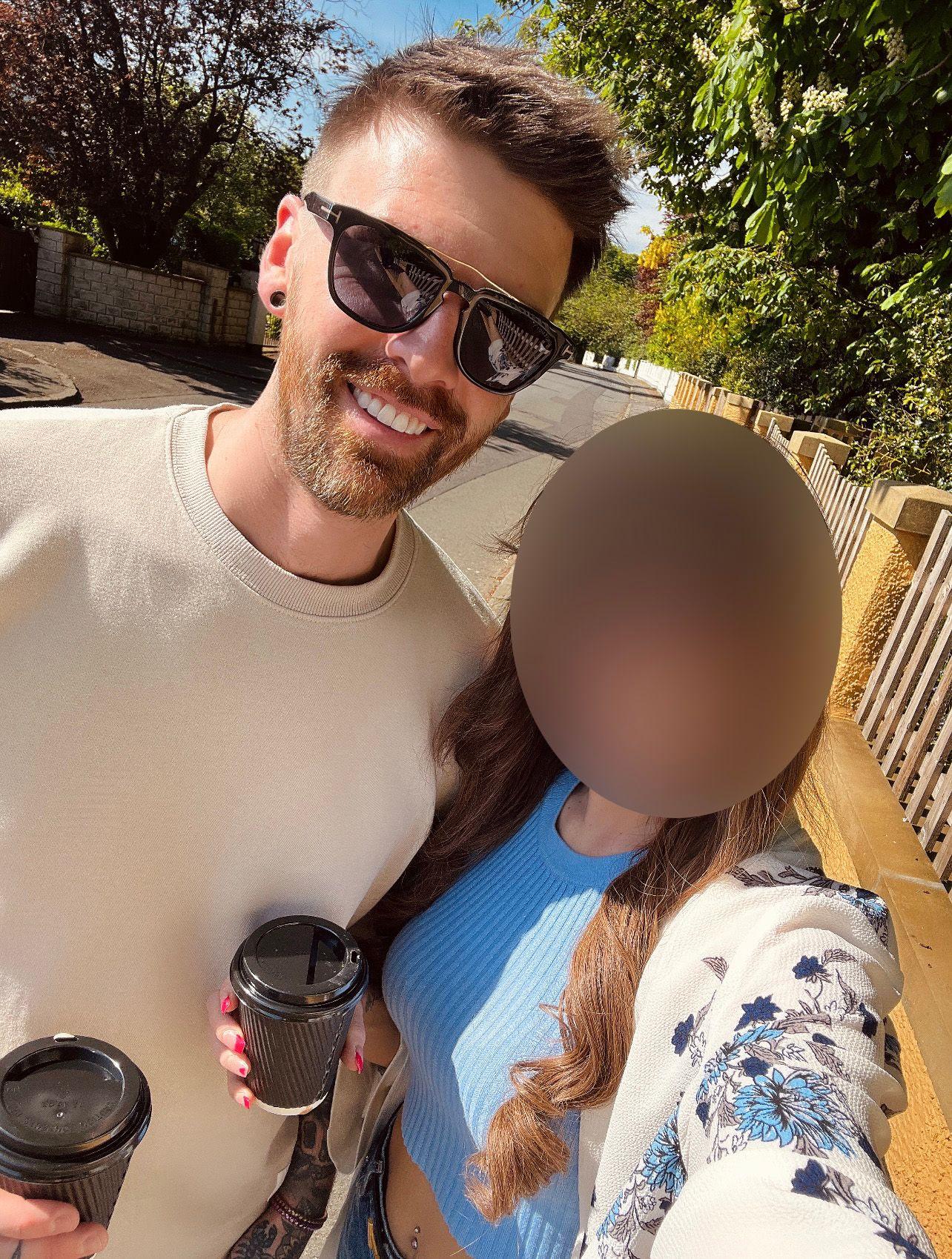 Un hombre y una mujer se toman una selfie al aire libre, sosteniendo dos cafés para llevar. El hombre tiene el pelo castaño y de punta, un jersey beige y gafas de sol oscuras. La mujer tiene el pelo largo y castaño, un top corto azul y un cárdigan, y su rostro aparece borroso.