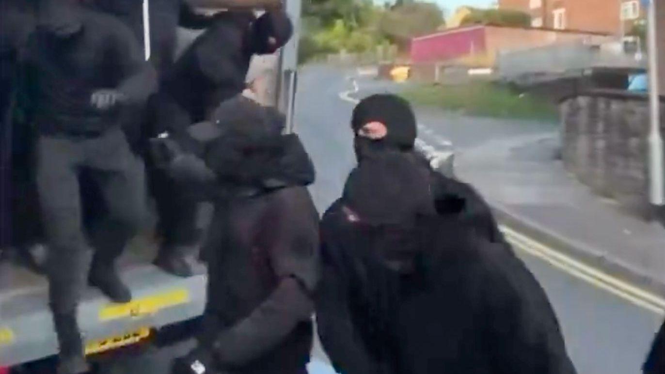 Hombres vestidos de negro y con pasamontañas bajando de la parte trasera de un camión.