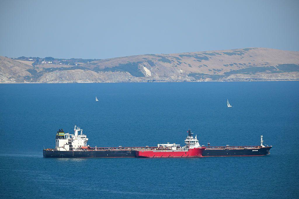 Un petrolero y un buque de apoyo en alta mar, frente a una costa rocosa, con dos veleros visibles al fondo.