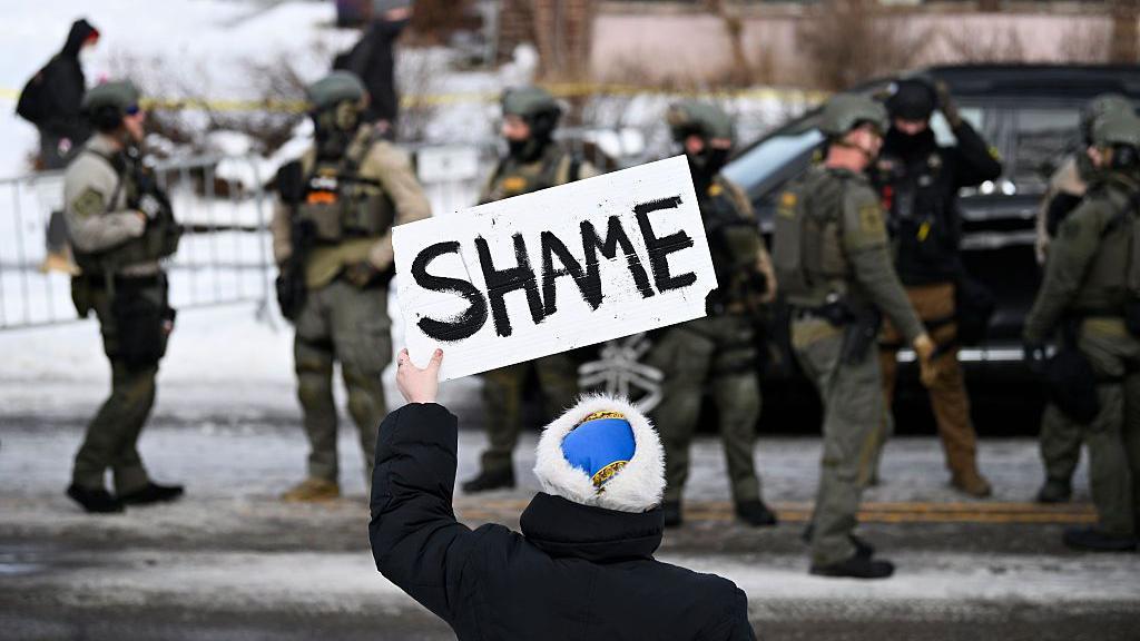 Una persona sostiene un cartel con la palabra "vergüenza" mientras se ven agentes de ICE durante operaciones federales de seguridad el 7 de enero de 2026 en Minneapolis, Minnesota.