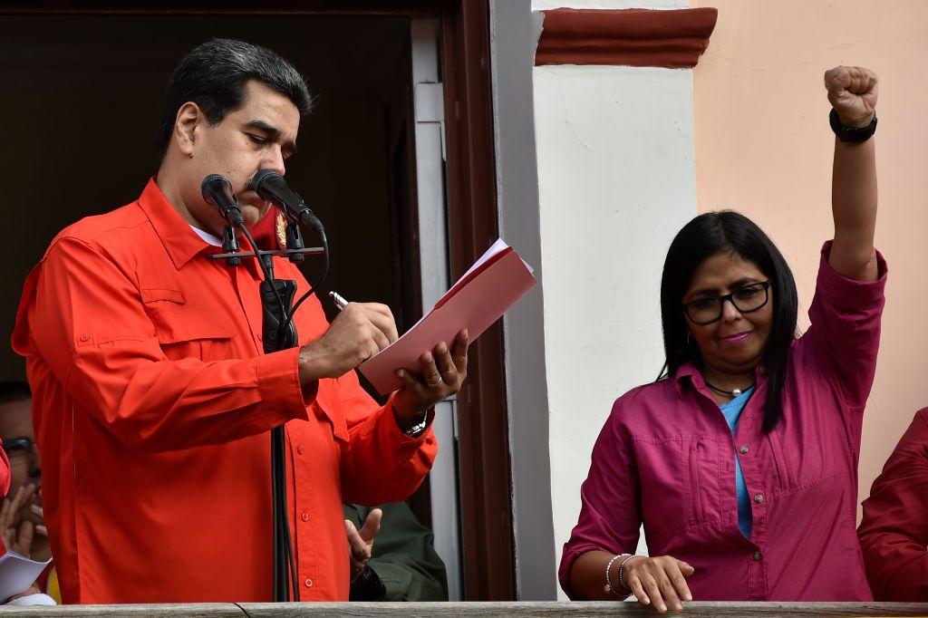 Nicolás Maduro viste una chaqueta roja mientras firma un docmento. A su lado está de pie Delcy Rodríguez levantando el puño izquierdo en alto.