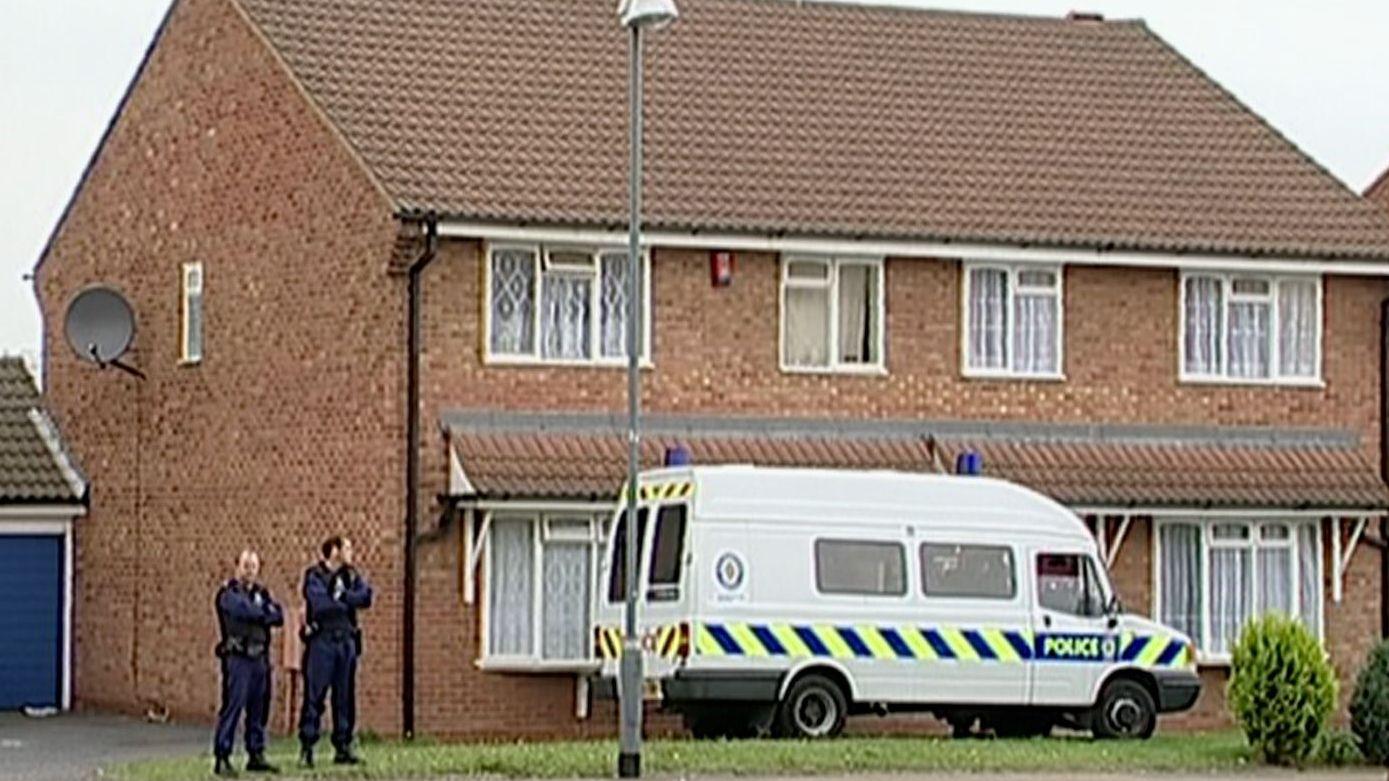 Imagen extraída de un vídeo que muestra una pequeña casa pareada de mediados del siglo XX en Birmingham, con visillos y una antena parabólica. Una furgoneta policial está aparcada en el exterior, bloqueando la puerta de entrada, y dos agentes de policía permanecen a un lado del edificio con los brazos cruzados.
