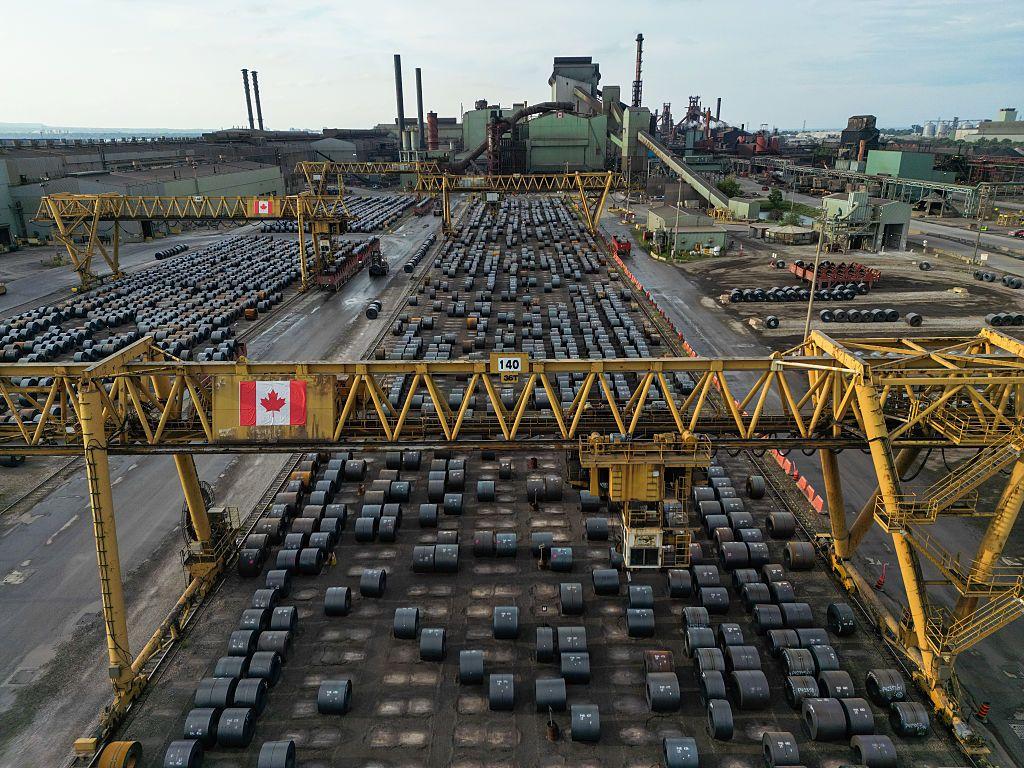 Una vista aérea en picado muestra hileras ordenadas de rollos de acero almacenados en el patio de una planta siderúrgica canadiense, con grandes grúas pórtico amarillas sobre la cabeza.