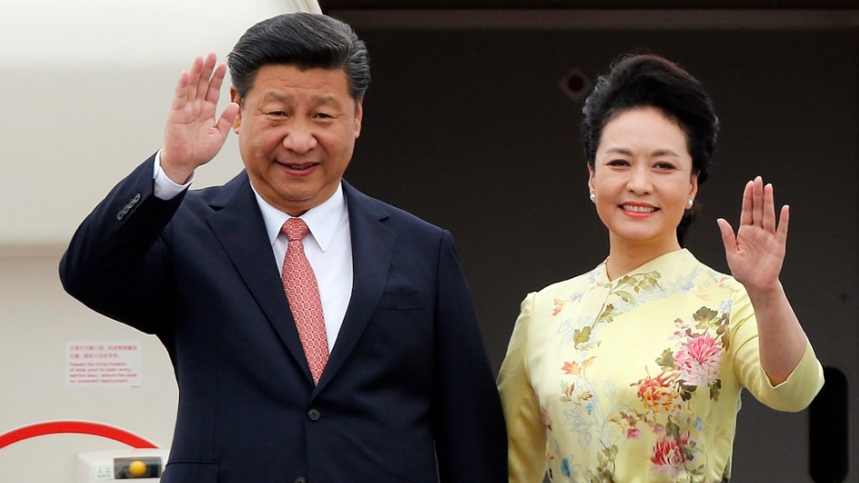 Peng Liyuan, con un vestido floral, y Xi Jinping, vestido con traje y corbata roja, saludan con la mano frente a la puerta abierta de un avión.