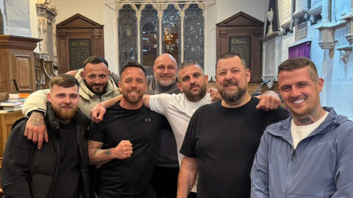 Un grupo de siete hombres sonrientes de pie en el pasillo de una iglesia con una vidriera detrás.
