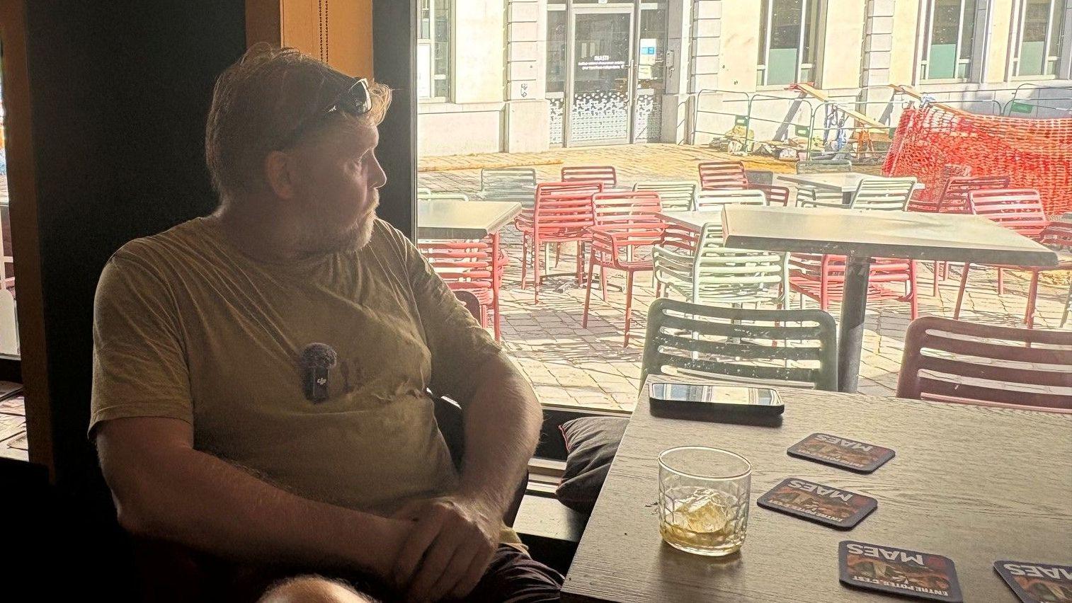Un hombre, pensativo, mira a través de la ventana de un café. Tiene una bebida sobre la mesa frente a él, y afuera, en la calle, hay sillas de colores.