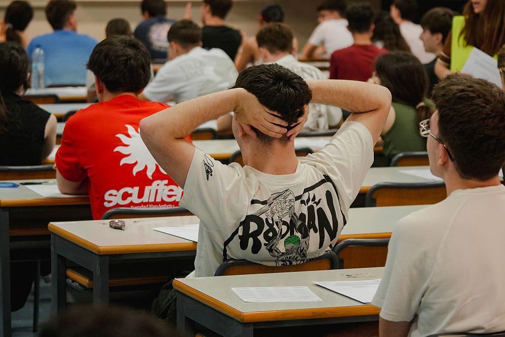 Un grupo de estudiantes sentados en un aula de Valencia, España. Se les ve de espaldas. La imagen la domina uno de ellos que se lleva las manos a la nuca. 