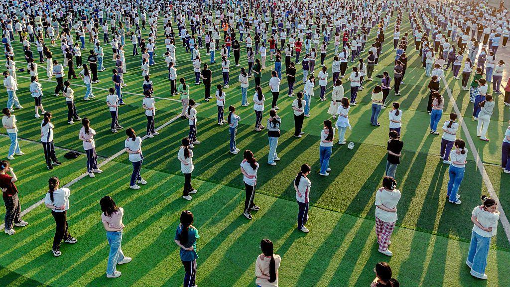 Cientos de estudiantes practican yoga en una cancha deportiva para aliviar el estrés antes del examen anual de ingreso a la universidad en China. Mantienen la misma distancia entre sí y visten ropa normal, no uniforme escolar.