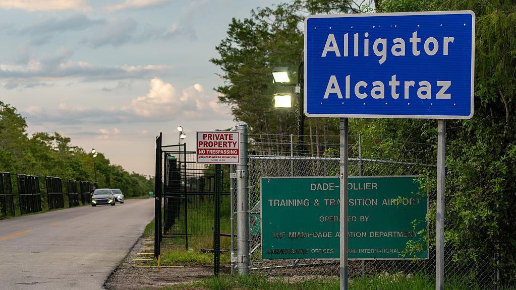 Un letrero azul que dice Alligator Alcatraz en la entrada de un camino, seguido de una larga carretera bordeada por una cerca metálica de seguridad. 