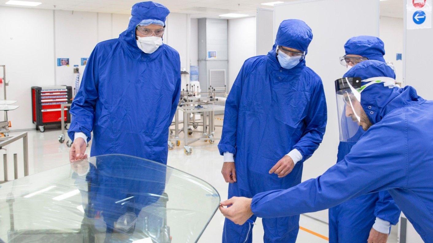 Cuatro trabajadores con monos azules inspeccionan la ventanilla de un avión. Llevan mascarillas y capuchas.