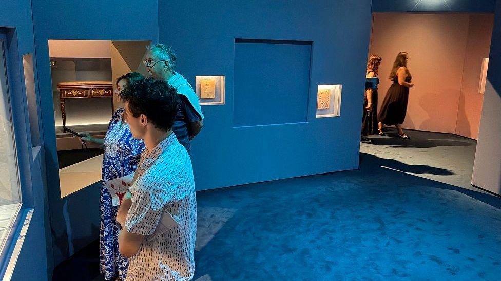 Varias personas observan una vitrina situada a la izquierda de una habitación con moqueta y paredes azules. Hay una pared con un cuadrado vacío.