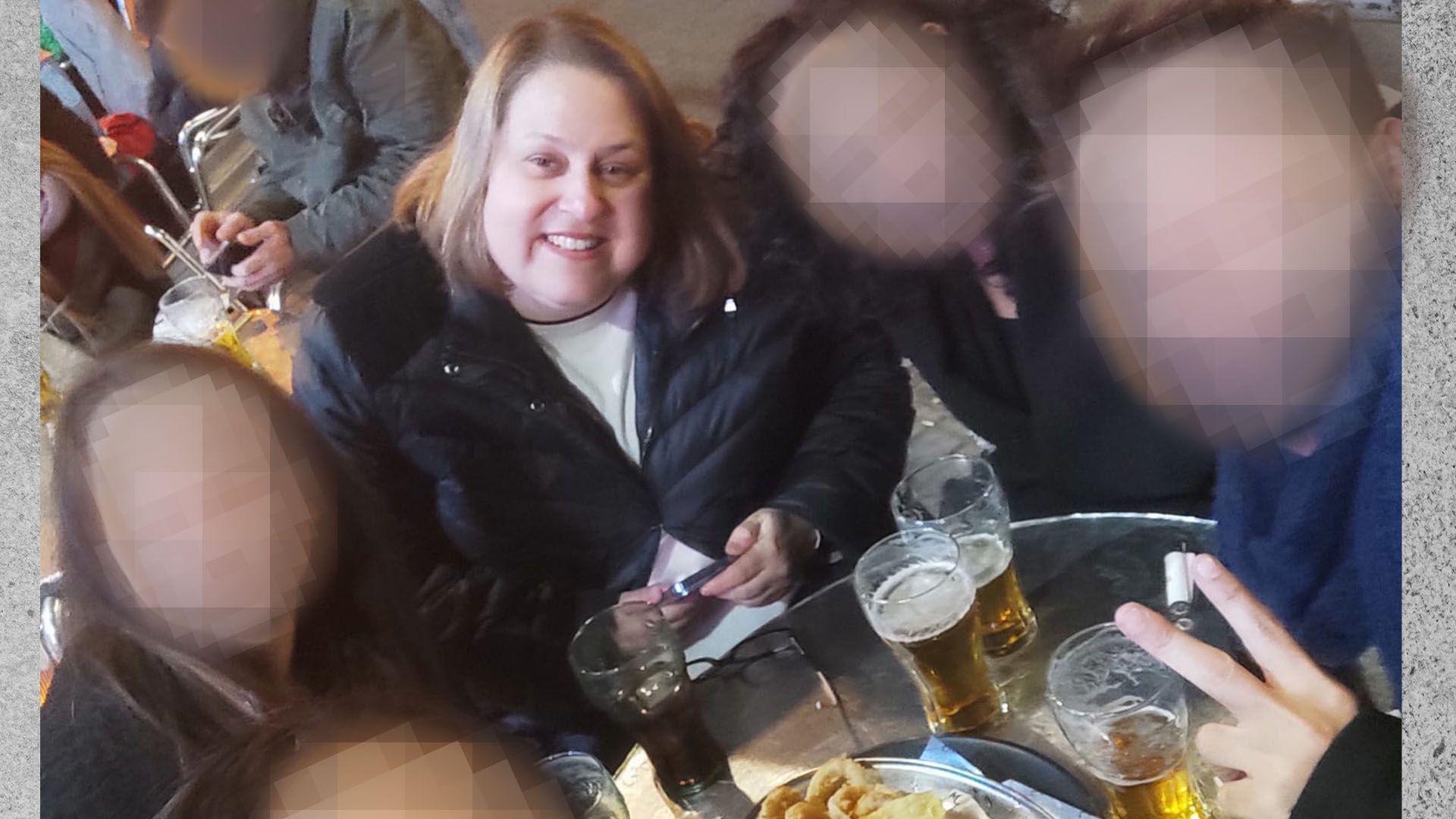 Una mujer de pelo rubio sonrie a la camara mientras toma un aperitivo con amigos. 