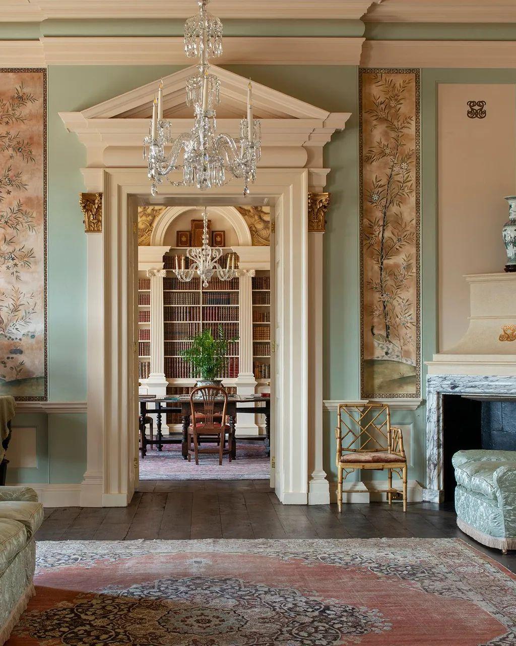 Fotografía a color tomada desde un salón en el que se ve una alfombra, una lámpara de cristal, una chimenea y un silla elegantes, así como el elaborado arco de la puerta, que deja ver al fondo una biblioteca, con otra lámpara de cristal, una mesa y sillas, y una planta.  