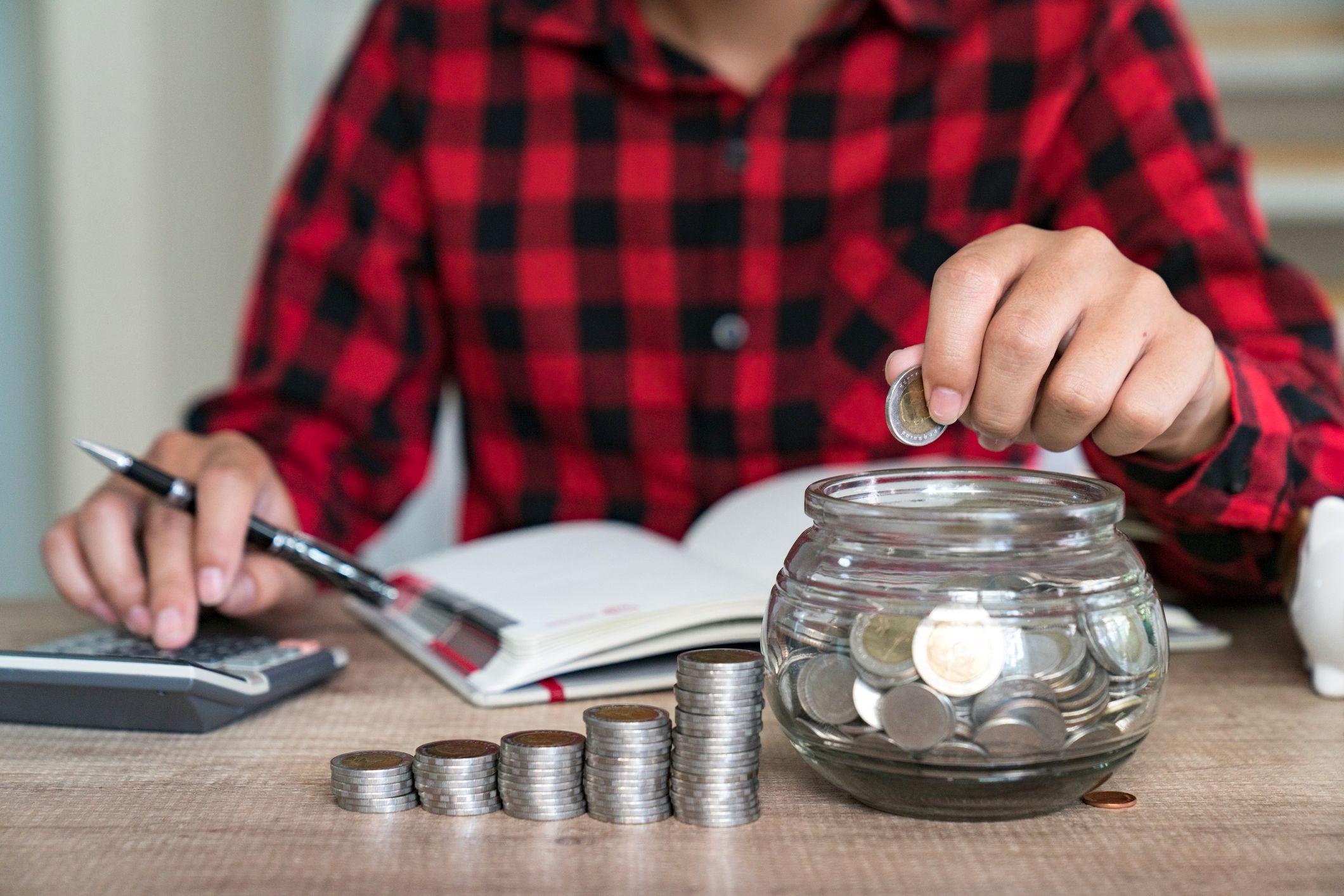 Una persona haciendo anotaciones en un cuaderno con unas pilas de monedas en frente y un recipiente de vidrio con monedas. Tiene además una calculadora