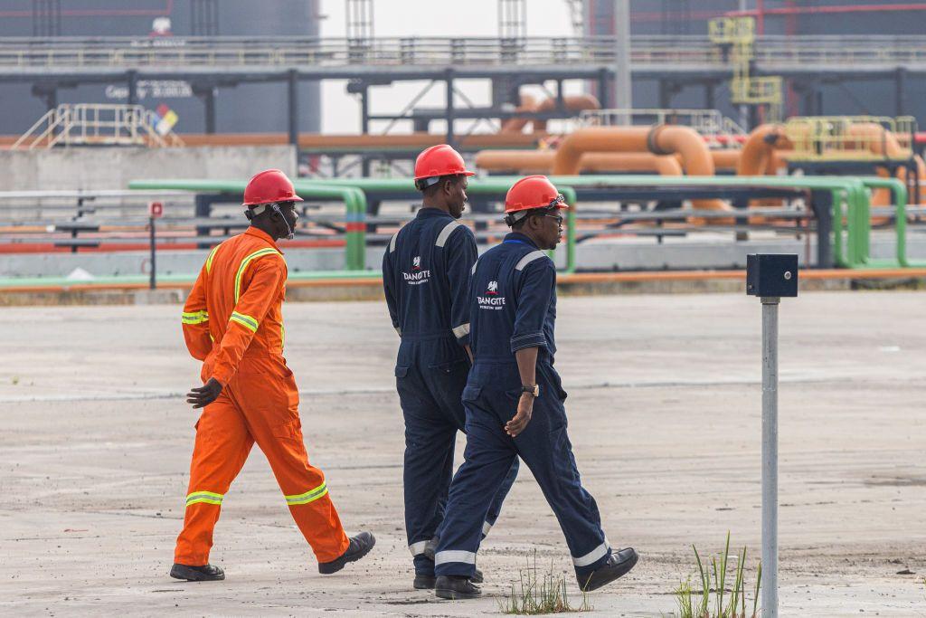 Trabajadores en la refinería de petróleo y planta de fertilizantes de Dangote Industries Ltd. Los tres hombres llevan cascos rojos. Dos visten monos de trabajo azul marino con el logo de Dangote, mientras que el tercero lleva un mono naranja. A lo lejos se observa una hilera de tuberías de petróleo de color naranja y verde.