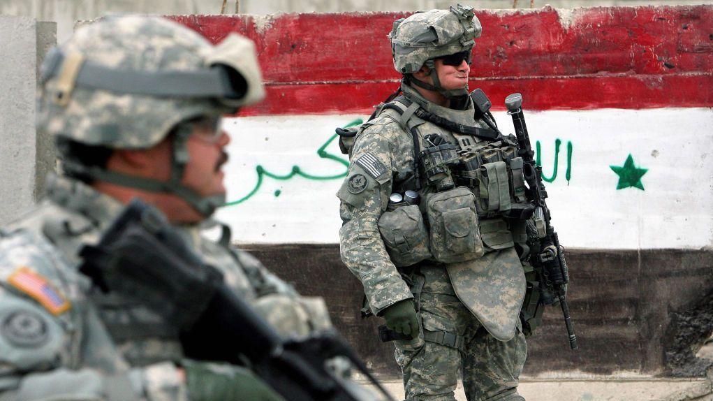 Dos soldados estadounidenses con equipo de combate permanecen de pie cerca de una barrera pintada con los colores de la bandera iraquí; uno de ellos está en primer plano y el otro sostiene un rifle al fondo.