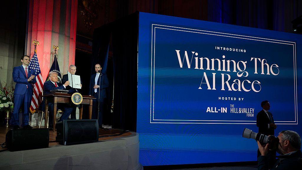 El presidente estadounidense Trump, flanqueado por varios miembros de su administración, se sienta detrás de un escritorio con el sello presidencial en el escenario durante un evento en 2025. A su derecha hay un enorme cartel azul con la inscripción "Ganando la carrera de la IA" en letras blancas. Sostiene una carpeta que contiene una orden ejecutiva que acaba de firmar sobre inteligencia artificial.