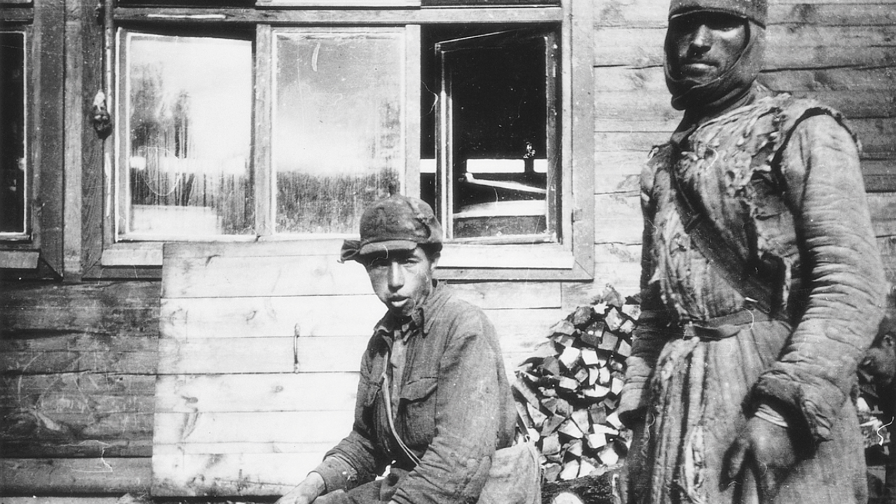 Una fotografía en blanco y negro tomada en Alderney muestra a dos prisioneros de guerra con chaquetas y gorros acolchados de algodón. El del centro está sentado, el de la derecha de pie, sosteniendo lo que parece ser una herramienta agrícola. Detrás de ellos se ve una cabaña de madera con las ventanas abiertas.