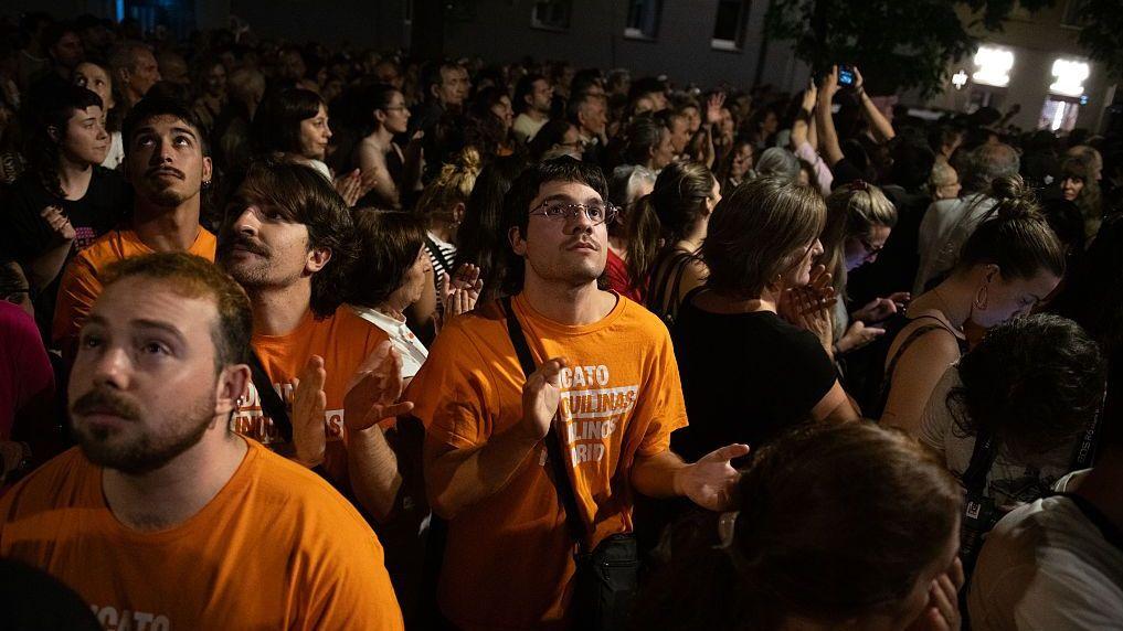 Manifestantes con camisetas naranjas se congregan en una calle oscura. 