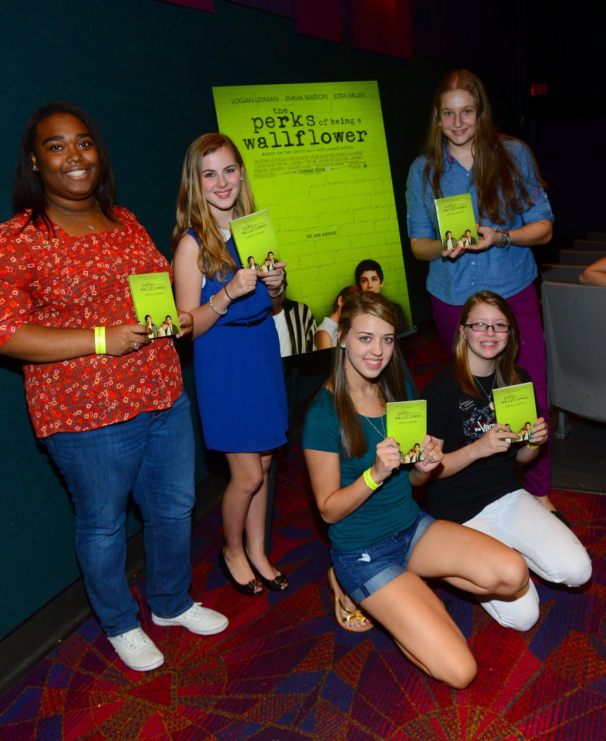Cinco adolescentes miran a la cámara mientras sostienen en sus manos una copia de "Las ventajas de ser invisible" alrededor de un póster de la película del mismo título.