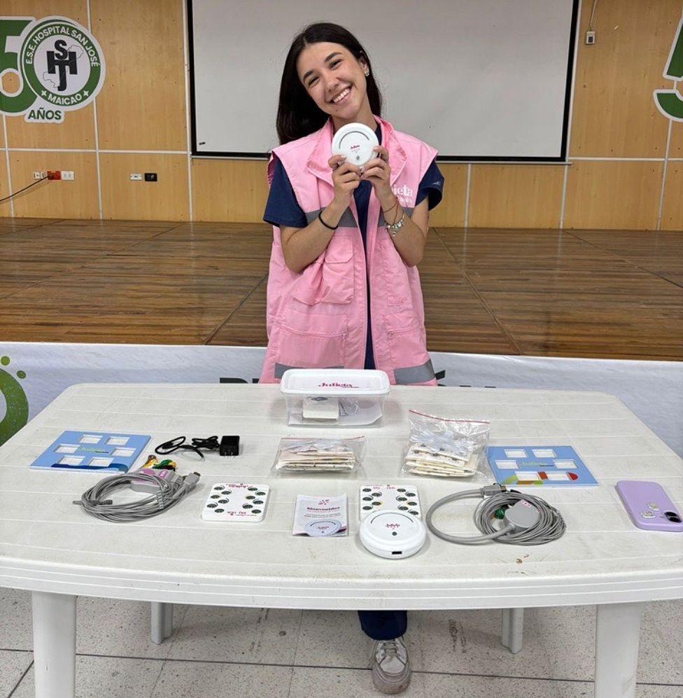 Chica sosteniendo el dispositivo con ambas manos, sonriendo, con pelo largo oscuro, una camisa azul oscura con un chaleco rosado encima, frente a una mesa blanca en la que se ven cables y otros artefactos. 