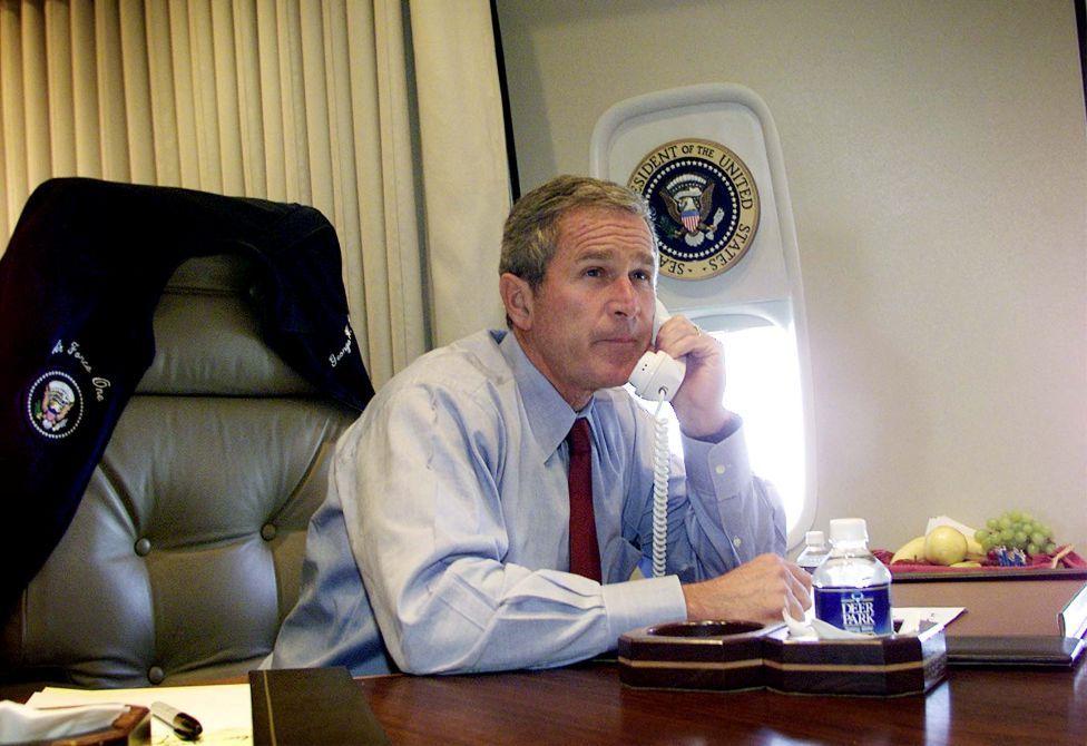 El presidente Bush está sentado en un escritorio del avión, sin chaqueta, la cual cuelga en el respaldo de su asiento. Viste una camisa azul claro y una corbata granate. Tiene un teléfono en la mano izquierda y parece estar conversando.