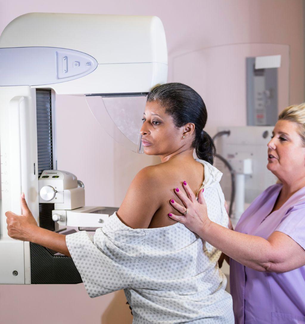 Una mujer de unos cuarenta años, vestida con una bata de hospital y sometiéndose a su mamografía anual. Recibe asistencia de una técnica, una mujer rubia que lleva uniforme sanitario.