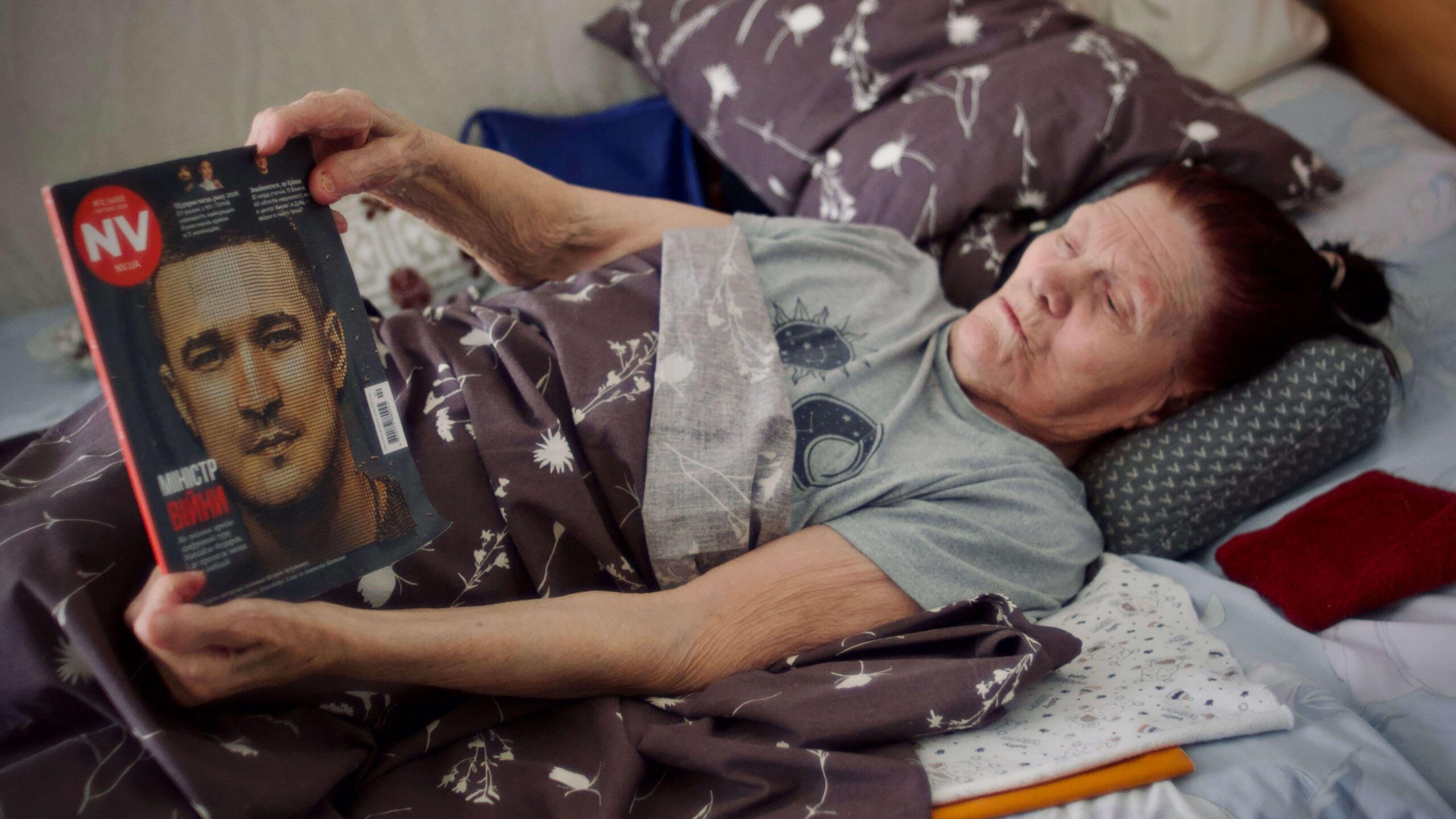 En la foto, Nadia está en la cama leyendo una revista. Lleva una camiseta gris y tiene el pelo oscuro.