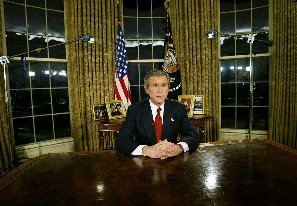 El expresidente estadounidense George W. Bush está sentado detrás de un escritorio en el Despacho Oval durante un discurso televisado, con banderas estadounidenses, grandes ventanales y equipos de iluminación de televisión visibles al fondo.