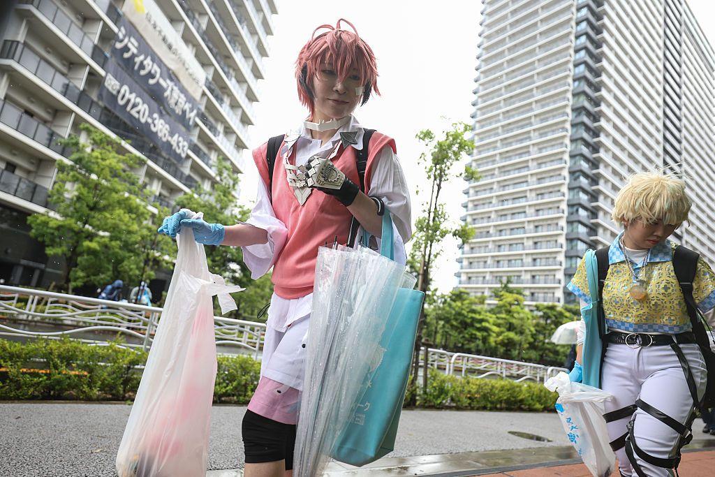 Dos Personas con disfraces, la de la izquierda color rosado y la de la derecha amarillo. Caminan en una calle con edificios y cargan bolsas con desperdicios tras una convención.