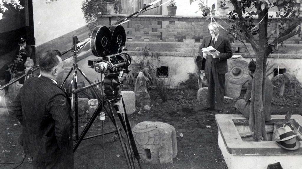 Foto en blanco y negro de Trotski con un libro en la mano en los jardínes de su casa en Coyoacán, Ciudad de México. Un camarógrafo lo filma.