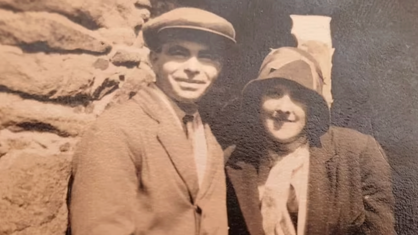 John y Phyllis Le Breton aparecen sonriendo en una antigua fotografía en sepia. Él lleva gorra y traje, y ella sombrero, abrigo y bufanda. Parecen estar de pie frente a un monumento de piedra.
